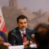 A photo-realistic image depicting a formal press conference in Iran. Several Iranian officials are seated at a table with microphones, addressing a group of journalists. The scene should convey a sense of seriousness and officialdom. The background should feature the Iranian flag and possibly a subtle depiction of a government building. Focus on the expressions of the officials, conveying confidence and resolve.