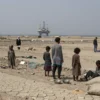 A stark, realistic scene depicting a severe famine in a region affected by the Ormuz crisis: emaciated families standing among barren, cracked fields under a bleak sky, empty bowls and broken grain sacks scattered around, distant silhouettes of a blocked shipping route or oil rigs on the horizon to hint at the Ormuz cause, with a somber, muted color palette emphasizing hardship and desperation