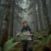 A man in hiking clothes stands in a dense, misty forest on Kamchatka. The forest is full of tall trees and ferns. The lighting is dim and atmospheric, suggesting a search and rescue operation. The man looks worried and is holding a map.