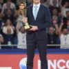 A sports commentator from Ivanovo, wearing a suit and tie, holding a trophy, standing on a podium with a cheering crowd in the background. The scene should be brightly lit and convey a sense of accomplishment.