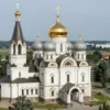 A large, ornate Russian Orthodox monastery under construction, located in the outskirts of Berlin. The monastery features multiple domes, golden crosses, and intricate architectural details. The scene should depict the monastery in a slightly unfinished state, with scaffolding visible. The surrounding landscape is a mix of green fields and trees.