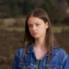 A realistic photo of a young woman, approximately 16 years old, with light brown hair and blue eyes, standing in a slightly overgrown field. She is wearing a denim jacket and jeans. The background shows a rural landscape with trees and a distant house. The overall mood is somber and slightly melancholic.