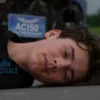 A solemn, realistic photograph of a young man, approximately 19 years old, lying on the road after a car accident. The scene should depict a damaged vehicle in the background, with a cloudy sky overhead. Focus on conveying a sense of tragedy and loss.