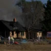 A photorealistic image depicting the aftermath of a house fire in a rural area. The focus should be on the charred remains of a residential building, with smoke still rising in the background. Emergency vehicles (fire trucks and ambulances) are present, indicating rescue efforts. The scene should convey a sense of tragedy and loss.
