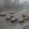 A wide shot of Ulianovsk city streets during road repair work. Construction vehicles, workers in safety vests, and road barriers are visible. In the background, street cleaners are actively sweeping the streets. The overall atmosphere is one of urgency and efficiency. Focus on the contrast between the ongoing repair and the quick cleanup.