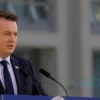 A photorealistic image of Bavarian Prime Minister Markus Söder speaking at a press conference. He is wearing a dark suit and tie, and standing behind a podium with the Bavarian state emblem. The background shows a blurred view of a modern building.