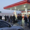 A long line of cars waiting at a gas station in Ulyanovsk, Russia. The gas station is modern and well-lit. People are standing in line, some looking impatient. The scene depicts the impact of gas station closures in other regions on Ulyanovsk residents.