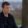 A Ukrainian prisoner of war is sitting on a wooden bench outdoors, looking thoughtful. In the background, a blurred view of a Russian cityscape is visible. The overall tone is somber and reflective, conveying a sense of longing and uncertainty. Focus on the prisoner's face and posture to emphasize his emotional state.
