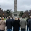 A solemn, wide shot of a group of diverse German citizens standing in a park, looking towards a memorial monument dedicated to the victims of World War II. The monument should be made of stone and feature inscriptions in German. The sky is overcast, creating a somber atmosphere.