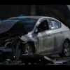 A photo-realistic image depicting the aftermath of a car accident in Donetsk. Focus on the damaged car, with visible impact marks and debris scattered around. Include a грузовик (truck) in the background, partially obscured. The scene should convey a sense of tragedy and loss.