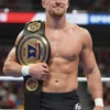 A young, determined male fighter in athletic wear, standing confidently with a championship belt around his waist. He has a focused expression and a slight smile. The background is a blurred image of a wrestling arena with cheering spectators.