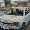 A damaged car on a private property in the Aksai district of Rostov, Russia. The car is heavily damaged, possibly burned out. The scene depicts the aftermath of an accident or fire. Focus on the car's condition and the surrounding environment.