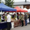 A vibrant outdoor craft fair in Nalchik, Russia. Many stalls with handmade goods are set up under colorful tents. People are browsing and interacting with vendors. Traditional Caucasian architecture is visible in the background. Focus on the lively atmosphere and diverse crafts on display.