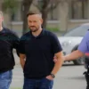 A photo-realistic image depicting two men being escorted by police officers in Rybinsk. The men should appear to be of similar age and build, wearing casual clothing. The police officers should be in uniform. The background should show a typical Rybinsk street scene with buildings and possibly some cars. Focus on a neutral, documentary style.