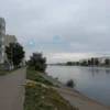 A quiet residential area in the Soviet district of Astrakhan, with a calm riverbank, trees, and a distant view of apartment buildings; the scene is peaceful and somber, with no people visible, evoking a sense of loss
