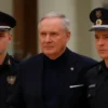 A serious-looking man in a dark suit being escorted by two uniformed Russian Ministry of Emergency Situations (MChS) officers. The setting is a modern, slightly blurred government building hallway. Focus on the man's face and the officers' posture.