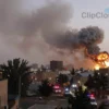 A wide shot of the city of Karaj, Iran, at dusk. Several large explosions are visible in the distance, with plumes of smoke rising into the sky. Buildings are silhouetted against the fading light. Emergency vehicles with flashing lights are approaching the scene. Focus on the urban landscape and the aftermath of the explosions, conveying a sense of chaos and urgency.