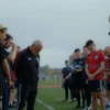 A photo-realistic image depicting a solemn farewell scene. A group of people, including coaches and players, are gathered around a figure (presumably Leonid Shchukin) who is standing with a slightly bowed head. The setting is a sports facility, possibly a stadium or training ground, with a slightly overcast sky in the background. The overall mood should be respectful and melancholic.