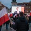 A dramatic scene of a large crowd of people protesting in a historic Polish city square at dusk, holding Polish flags, facing a large screen displaying a silhouette of a politician, angry and determined atmosphere, cobblestone pavement, classical European architecture in the background, cinematic lighting