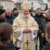 A medium shot of Bishop Alexander being greeted by people in Barysh. The bishop is wearing a religious robe and a pectoral cross. The people are dressed in everyday clothing and are smiling. The background shows a church and a town square.