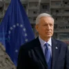 A photo-realistic image depicting the UN Secretary-General standing in front of the Venezuelan flag, with a backdrop showing the aftermath of an earthquake – damaged buildings and rescue workers. The scene should convey a sense of humanitarian aid and support.