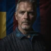 A serious and contemplative portrait of Vance, a middle-aged man with a thoughtful expression, standing in a dimly lit room with a Ukrainian flag subtly visible in the background. The lighting should highlight his facial features, emphasizing the gravity of his expression.