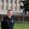 A professional security guard in a dark uniform standing confidently in front of a modern government building with the Russian flag, symbolizing official regulation and oversight of private security schools, clear daylight, sharp focus, realistic details