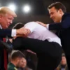 Donald Trump and Emmanuel Macron engaged in a friendly wrestling match on a brightly lit stage in a grand hall. The scene should depict a moment of playful competition, with both leaders smiling and showing effort. The background should feature a blurred audience and official flags of the USA and France. Focus on capturing the physical interaction and expressions of the two men.