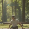 A person wearing headphones is sitting on a park bench, listening to music. The scene is bright and sunny, with trees and green grass in the background. The person is looking relaxed and enjoying the music.