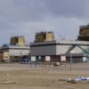 A photorealistic image depicting the Fukushima Daiichi Nuclear Power Plant in Japan, with visible damage from the recent earthquake and tsunami. Show damaged reactor buildings, debris scattered around, and a cloudy sky. Focus on the scale of the devastation and the power plant's location near the coast.