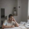 A young man, looking concerned, is sitting at a desk with a laptop. The laptop screen shows a phone conversation with a blurred figure. Scattered papers and a coffee cup are on the desk. The background is a simple office setting.