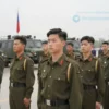 A group of young men in military uniforms standing in formation on a parade ground. They are looking forward with determined expressions. In the background, there are military vehicles and a flag. The sky is overcast.
