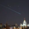 A wide shot of a nighttime cityscape of Moscow with a clear night sky. Several bright streaks of light representing intercepted drones are visible in the sky above the city. Military aircraft are flying in formation, positioned to defend the city. The city lights are visible below, creating a contrast with the dark sky and the streaks of light.