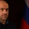 A serious-looking Nikol Pashinyan standing in a dimly lit room, looking concerned. In the background, a blurred image of the Russian flag is visible. The overall mood should be somber and reflective.