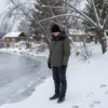 A 16-year-old boy standing near a frozen pond in a rural village in Krasnoyarsk Krai, surrounded by snow-covered trees and a quiet winter landscape, with faint footprints leading to the edge of the ice, solemn and natural atmosphere