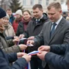 A group of people, likely officials from Smolensk UFSIN, are handing out small Russian tricolor ribbons to citizens. The scene takes place outdoors, possibly in a public square or during an event. Focus on the interaction between the officials and the people receiving the ribbons. The atmosphere should be positive and celebratory.