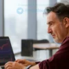 A photo-realistic image of a man in his 50s, wearing a casual shirt, looking thoughtfully at a laptop. The laptop screen displays stock market data and charts. The background is a modern, minimalist office space with large windows showing a city skyline.