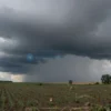 A dramatic sky over a rural landscape in the Voronezh region, Russia. Dark, stormy clouds are gathering, with visible hail falling from the sky. Fields of crops are visible below, with some areas showing damage from the hail. A few scattered trees are silhouetted against the sky.