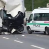 A damaged Hino truck and a damaged GAZelle van after a collision on a road. Debris is scattered around. Emergency vehicles (police car, ambulance) are present. The scene is outdoors during daylight. Focus on the vehicles and the immediate aftermath of the accident.