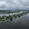 Aerial view of a vast flooded area in Russia, showing submerged fields, roads, and small villages with houses partially underwater. The sky is overcast with dark clouds, indicating recent heavy rainfall. The water reflects the surrounding landscape, creating a sense of depth and scale.