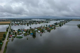 Aerial view of a vast flooded area in Russia, showing submerged fields, roads, and small villages with houses partially underwater. The sky is overcast with dark clouds, indicating recent heavy rainfall. The water reflects the surrounding landscape, creating a sense of depth and scale.