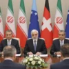 A wide shot of the Iranian delegation at the negotiations in Switzerland. The delegation consists of several men in formal attire, seated around a table with other representatives. The setting is a formal conference room with flags of various countries in the background. Focus on the serious expressions of the delegates and the overall atmosphere of the meeting.