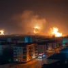 A panoramic view of Kharkiv city at night, with several explosions illuminating the sky and causing smoke plumes. Buildings in the city center are visible, with some showing signs of damage. Emergency vehicles with flashing lights are present on the streets. The overall mood is somber and chaotic, reflecting the intensity of the situation. Focus on the urban landscape and the effects of the explosions.