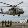 A military helicopter hovering over a remote island in the Pacific Ocean. In the foreground, a small group of soldiers are securing the area. In the background, a destroyed drug lab is visible. The scene is overcast and dramatic.