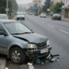 A damaged car on a road with debris, showing the aftermath of a collision. The road is asphalt, with visible lane markings. The background shows a blurred cityscape of Yekaterinburg, Russia, with buildings and trees. Focus on the damaged vehicle and the immediate surroundings.