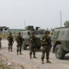 A panoramic view of a military checkpoint in a rural landscape. Soldiers in camouflage uniforms stand guard near armored vehicles. In the background, a damaged building suggests recent conflict. The sky is overcast, creating a somber atmosphere. Focus on realism and detail in the soldiers' equipment and the environment.