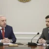 A formal meeting between Alexander Lukashenko and Volodymyr Zelenskyy. Both are seated at a large table in a neutral, modern conference room. The atmosphere is serious and diplomatic. Focus on their faces and body language, conveying a sense of negotiation and potential tension.