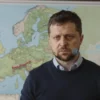 A serious and concerned-looking person, possibly a politician or diplomat, standing in front of a map of Europe with a red line indicating a potential conflict zone in Russia. The background should be slightly blurred to focus on the person and the map. The overall tone should be somber and reflective.
