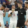 A soccer referee is shown ejecting three soccer players during a World Cup opening match. The referee is pointing towards the players with a stern expression. The three players are visibly upset, with body language indicating frustration and disagreement. The background shows a blurred stadium with fans. Focus on the referee and the players' reactions.