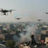 Aerial view of a cityscape with multiple drones flying in the sky, some of them being shot down by anti-aircraft systems. The scene should depict a chaotic and tense atmosphere with smoke and debris falling from the sky. The city below should show signs of damage and emergency response activities.